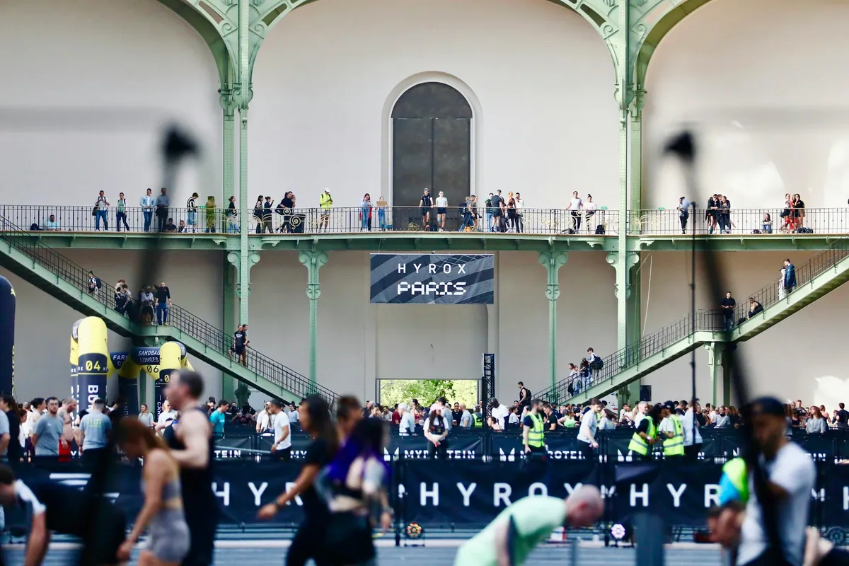 Athletes compete at a HYROX race in a large hall, with spectators on the balconies