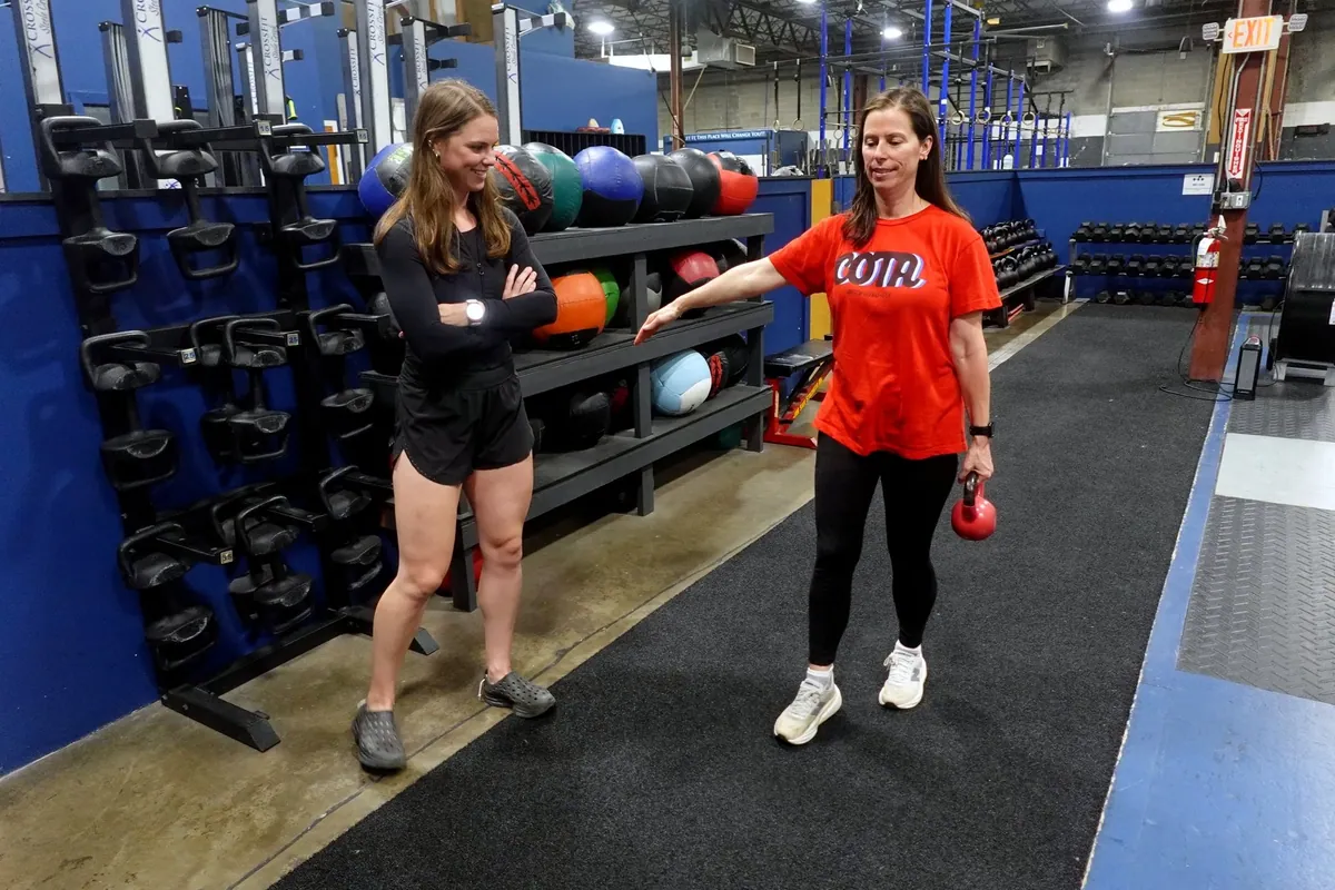A physical therapist coaches an athlete through a kettlebell carry in the gym