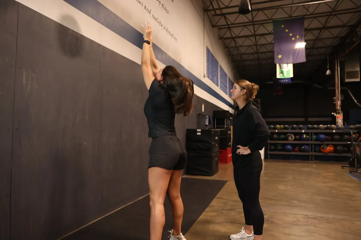 An athlete reaches up to a wall ball target while Kiley Robb, PT, DPT, watches