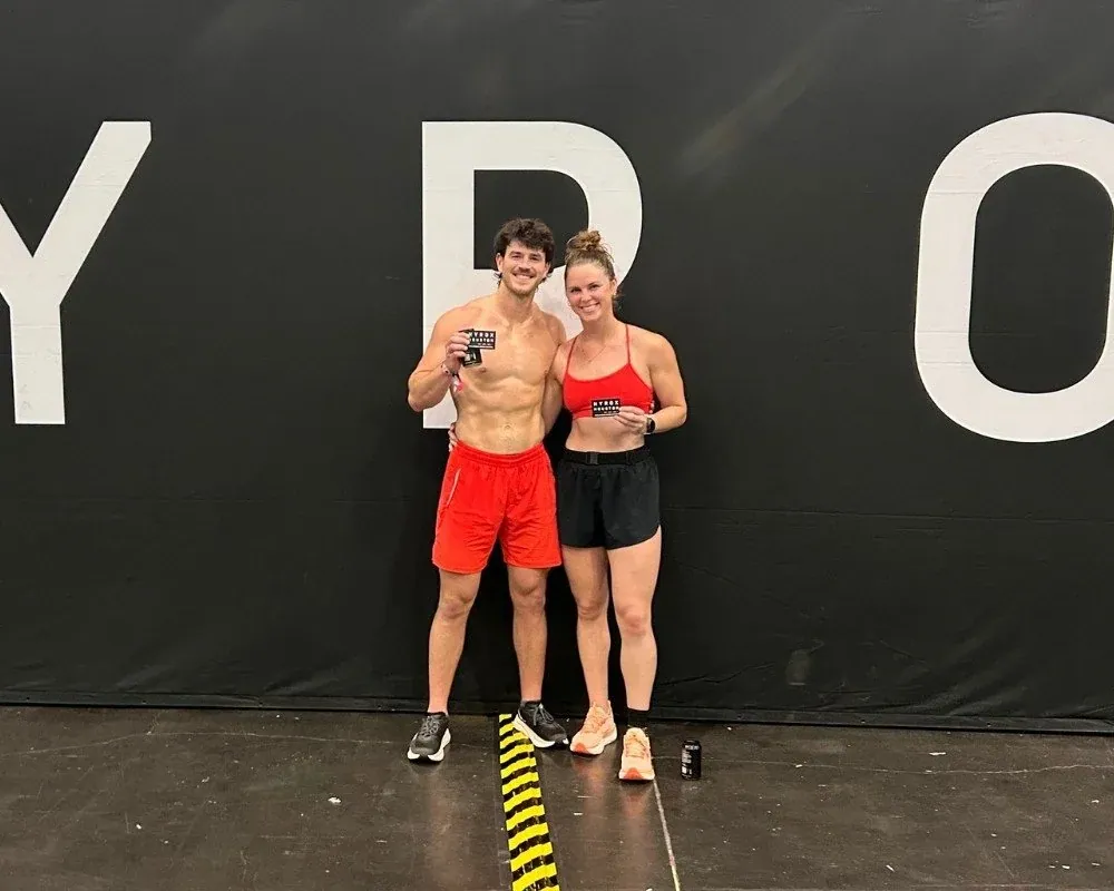 Two HYROX athletes hold their finisher patches in front of a HYROX wall
